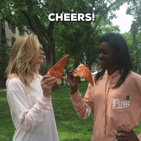 Ad gif. Two college-aged girls wearing Victoria's Secret PINK brand hoodies raise a slice of cheese pizza to each other and then take a bite with smiles.