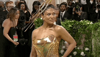Met Gala 2024 gif. Closeup of Sabrina Harrison wearing a smooth gold-colored fitted bodice with a scooped sweetheart necklace that is filled with a melting Dali-esque clock down the front. She's wearing a gold bedazzled fascinator in the style of a clock face with rods extending from its center with numbers at the endpoints. 