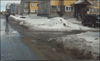 Video gif. We drive through a snowy neighborhood as a mailman walks on the slide of the road. The mailman walks into someone’s snow covered lawn and then falls into a waist high hole that was covered in snow.