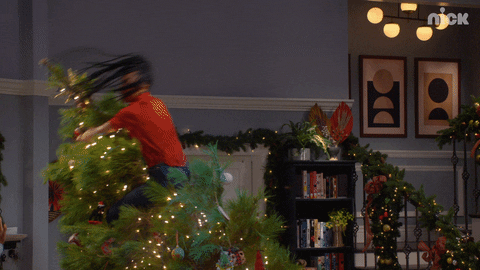 TV gif. In a scene from That Girl Lay Lay, a person with long dreadlocks clings to the top of a decorated Christmas tree as it starts to tip over.