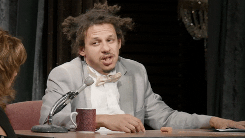 TV gif. Eric Andre on The Eric Andre Show sits at his desk looking over at a woman who's talking with a bored expression. He chews gum with his mouth open.