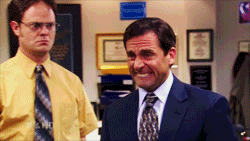 The Office gif. Zoom in on Steve Carell as Michael who is standing next to Rainn Wilson as Dwight. Michael cringes nervously, biting his lower lip with all his teeth. His face is bright red.