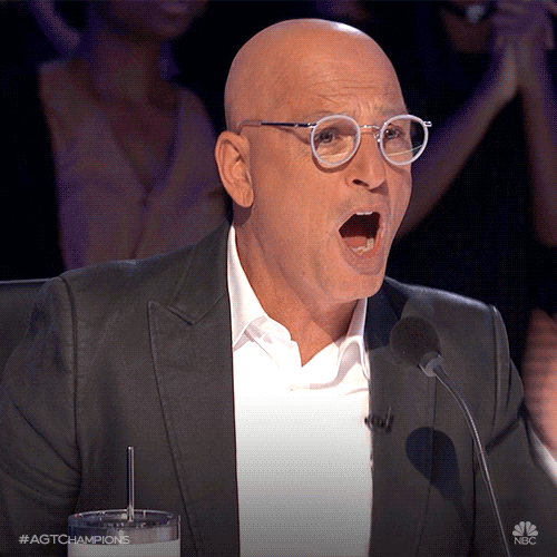 TV gif. Howie Mandel on America's Got Talent sits at a microphone and opens his mouth wide in shock as he covers it with his hand.