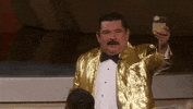 Oscars 2024 GIF. Guillermo holds up a tequila margarita and faces the audience. He's wearing a gold sequined tuxedo jacket. He cheers the crowd before clinking glasses with an audience member.