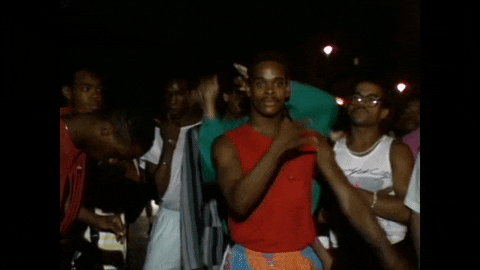 Video gif. Man in a vibrant red tank top confidently vogues in a crowd, getting down low as another man in a green shirt vogues on top of his head.