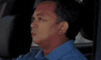 Video gif. A man driving a car slowly turns towards us and then smiles widely and gives an enthusiastic thumbs up.