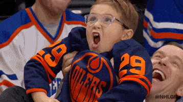 Sports gif. A young boy wearing glasses and an Edmonton Oilers jersey is lifted up in the air at a game. He screams in excitement and waves both hands as he celebrates the score.