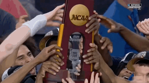 Sports gif. The UConn men's basketball team celebrate their win at the NCAA championship, crowding together as they smile and hold up their trophy.