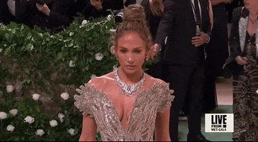 Met Gala 2024 gif. Closeup of Jennifer Lopez as she places a hand on the hip and poses with a confident expression. She's wearing a near-see-through Schiaparelli sleeveless gown with a plunging v-neckline that culminates in a pattern that flares out from her shoulders like butterfly wings. She's wearing a prominent diamond necklace, also resembling feathers. 