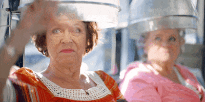 Video gif. Two older women sit under bonnet hair dryers and give emphatic thumbs down with faces of disgust.