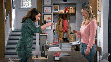 TV gif. Abby Elliott as Rebecca and Fran Drescher as Debbie from Indebted cheers their shot glasses together at a kitchen table before throwing back the alcohol and smiling.