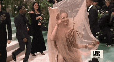 Met Gala 2024 gif. Lana del Rey wearing an Alexander McQueen with a veil held up by antlers, smiles broadly and waves for the cameras, then suddenly turns and shifts into a moody pose.