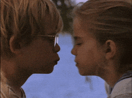 Movie gif. Anna Chlumsky as Vada and Macaulay Caulkin as Thomas in My Girl are in front of a lake and share a small smooch before separating with surprise on their faces.