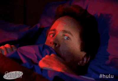Seinfeld gif. Jerry Seinfeld as himself lies terrified in bed as he holds the blankets up tight to his face. He glances worried around the room.