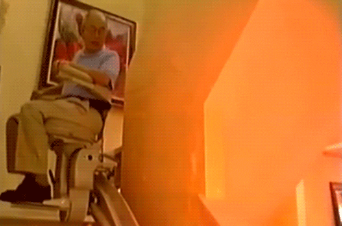 Video gif. A man is riding on a personal stair lift in his home but the stair lift slowly descends him into a fiery pit.