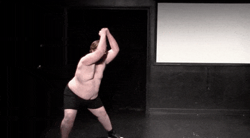 Video gif. A large man in black shorts without a shirt does a pelvic thrust, then launches into a somersault onto a bed of tacks scattered on the ground. 