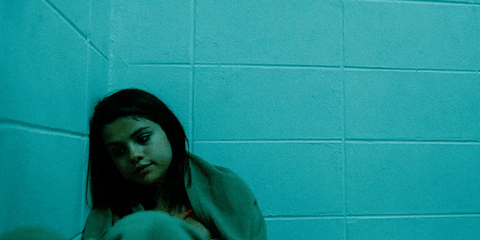 Movie gif. Lonely Selena Gomez as Faith in Spring Breakers leans against a cinderblock wall, looking sad and tired.