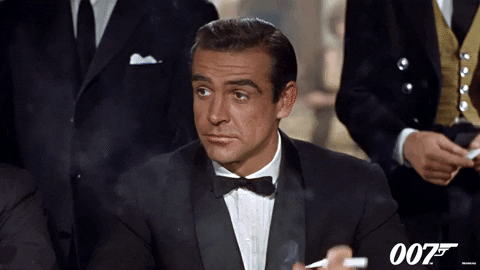Movie gif. Sean Connery as James Bond in 007 raises his hand, holding a cigarette with an expression that says, “count me in.”