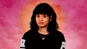 Celebrity gif. Kehlani stands in a black outfit in front of a mystical, cloudy background that has a radial gradient from orange to bright pink. She smacks on her gum while she widens her eyes suspiciously at us and looks away.