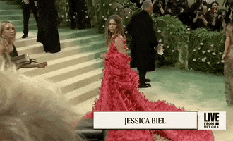 Met Gala 2024 gif. Side angle wide shot of Jessica Biel posing for pictures, wearing a hot pink Tamara Ralph Couture full-length gown with a deep-v neckline. The full billowing skirt flares out to a dramatic train. She wears a billowing shrug that matches the pattern of her dress around her back and arms. 