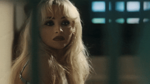 Music video gif from Sabrina Carpenter's music video for "Please Please Please." She sits in a jail cell behind bars, smiling and scrunching up her face, then stands up.