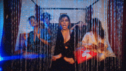 Music video gif. Dinah Jane in Heard It All Before sits at the foot of a bed, dancing in unison with her background dancers behind a beaded curtain. 