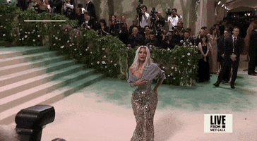 Met Gala 2024 gif. Zoom in on Kim Kardashian wearing a silver Maison Margiela gown and gray cardigan bolero, posing for the cameras, holding her sweater in place.