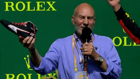 Sports gif. Patrick Stewart is on stage with Daniel Ricciardo at a Formula One event. Patrick is holding a boot filled with a drink and he chugs the whole thing, smiling when he's done and Daniel cracks up and pats him on the back.