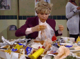 Video gif. Woman wearing a white plastic bib eats her way through a hoard of plates, food wrappers, and condiment bottles. She grabs at food and shoves it into her food covered mouth with determined intensity. 