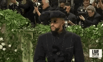 Met Gala 2024 gif. Closeup of the top of Bad Bunny's all navy-washed Margiela smoking jacket with decorative white stitching worn over a black satin corset. He's wearing an oversized puffy beret with floral details and diamond-shaped dark gold-framed sunglasses.