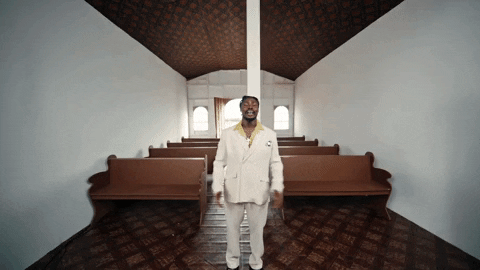Music video gif. Fireboy DML is standing in a church for his music video for Bandana. He has his hands clasped in front of him as he sings and the camera zooms in closer.