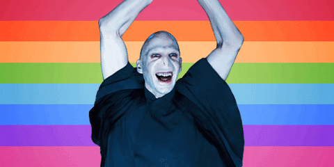Digital art gif. Mirrored shifting photo of Ralph Fiennes as Lord Voldemort grinning, holding his bare arms up in the air, in front of a rainbow-striped background.