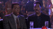 Movie gif. Eddie Murphy as Prince Akeem and Arsenio Hall as Semmi sit in a nightclub staring at something with shock and open mouths.