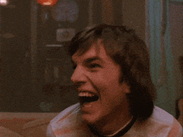TV gif. Ashton Kutcher as Kelso in That's 70s Show. He's sitting in the foggy garage and rocks back and forth with laughter and a dopey smile fills his face.