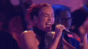 ACM Awards gif. Dua Lipa grips standing mic while singing with eyes closed and teeth showing, she raises her index.