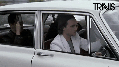 Music video gif. Fran Healy of Travis drives a vintage white car with cool focus as trees whiz by in the background. Text, "Is it Friday yet?