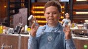 Reality TV gif. Young boy on Master Chef Junior Australia has a nervous smile on his face and crosses both of his fingers.