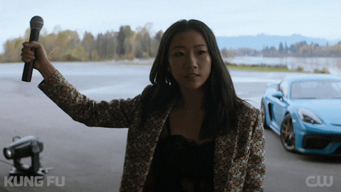 TV gif. Olivia Liang as Nicky Shen on Kung Fu has a serious expression on her face as she drops a microphone out of her hand. Behind her is a blue sports car.