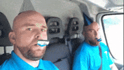 Video gif. In the backseat of a truck, two bald men with similar beards, bright blue shirts, and party hats blow into birthday noisemakers and stare directly at us.