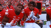 Sports gif. Three football players on the Maryland Terrapins sit on the sidelines. They pose for us, holding out peace signs, holding their hands in a praying position. One of them hugs the other and holds his thumbs up.