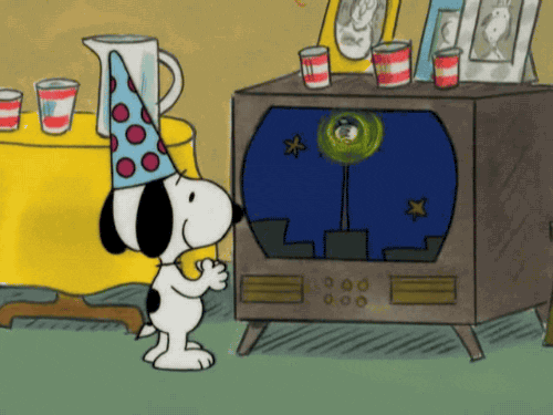 Cartoon gif. Snoopy wears a party hat and rubs his hands together as he watches the glowing New Year's Eve Ball on TV.