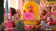 Reality TV gif. A person dressed in yellow with large yellow wig and pink gloves sits on a yellow couch with children dressed in extravagant outfits. The person holds up a pink sign with a large no written on it, drops it, then shrugs.