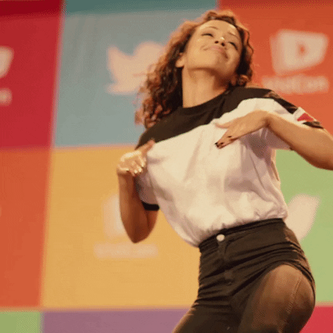 Celebrity gif. Wearing a white t-shirt with a black stripe along the shoulders and high-waisted pants, Liza Koshy dances cheerfully in front of a colorful wall of sponsorship logos.