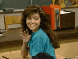 TV gif. Tiffani Thiessen as Kelly in Saved By The Bell. She's sitting in the front row of class and she turns around to smile cutely at someone and wave.