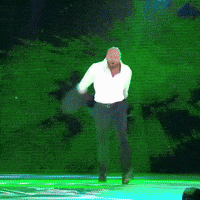 Video gif. Man takes off his suit jacket and loosens his collared shirt, running across a stage to meet a waiting opponent in a boxing rink.