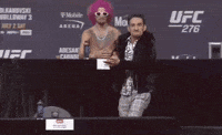 Sports gif. Wearing a furry black jacket and a complicated black-and-white shirt, a very intense Max Holloway claps, then flexes as he yells: Text, "Let's go!" Finally, he slaps his chest and holds up his arms.
