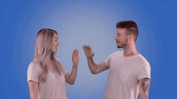 Video gif. A man and a woman in front of a blue backdrop smile at each other and high five, then turn and smile atus. The text, “teamwork!” appears above them when they high five.