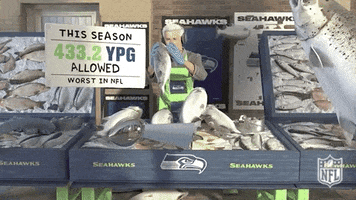 Sports gif. Pete Carroll is manning a fish stall in the NFL video game. He's trying to dodge the massive amount of fish being thrown at him but is unsuccessful in his efforts.