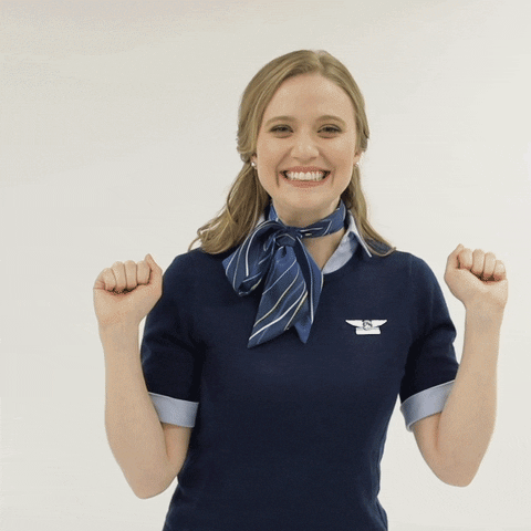 Video gif. An Alaska Airlines flight attendant with curly blonde hair smiles excitedly as she pumps her fists by her shoulders and dances in a circle.