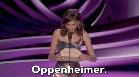 Oscars 2024 gif. Zendaya wears a glimmering, purple holographic dress that coordinates with the royal purple backdrop. She reads off of the envelope and announces Oppenheimer for Cinematography. 